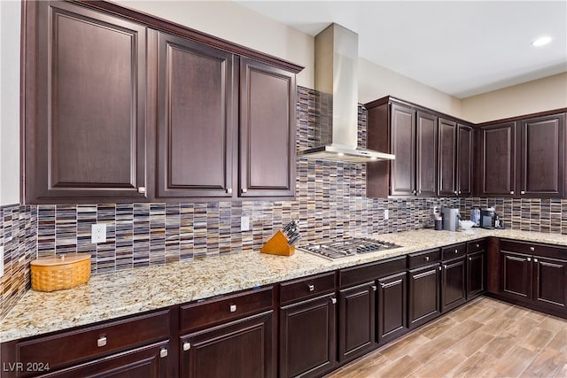 kitchen with wall chimney range hood, light hardwood / wood-style floors, decorative backsplash, stainless steel gas cooktop, and dark brown cabinets