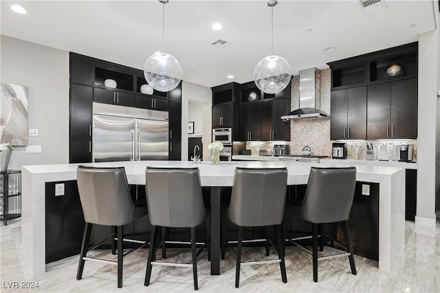 kitchen featuring wall chimney exhaust hood, decorative light fixtures, a large island with sink, and appliances with stainless steel finishes