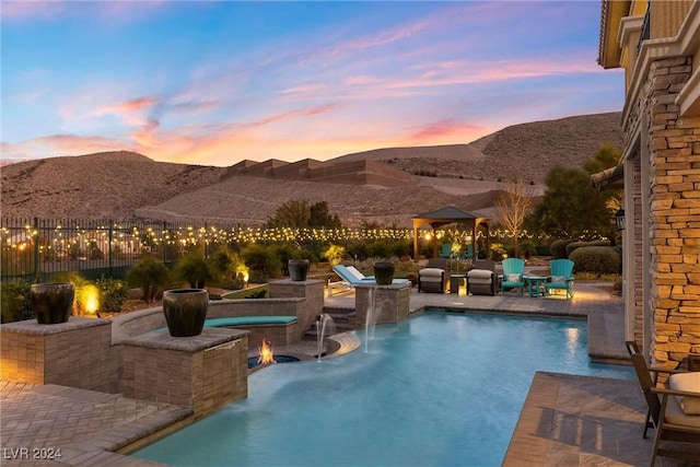 pool at dusk with a jacuzzi, pool water feature, an outdoor fire pit, a patio area, and a mountain view