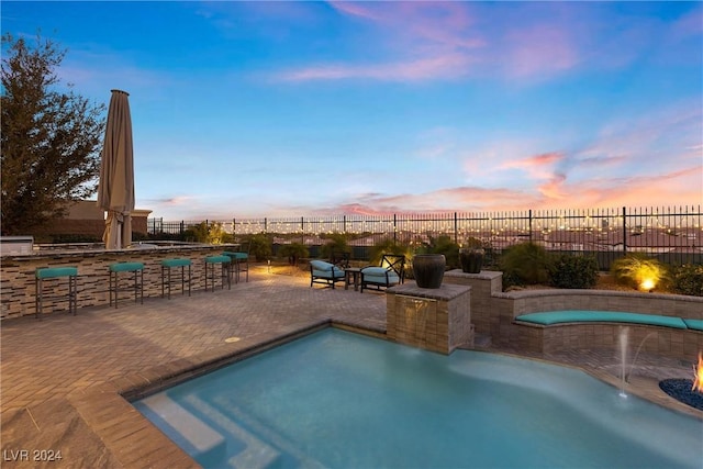 pool at dusk featuring pool water feature, an outdoor bar, and a patio area