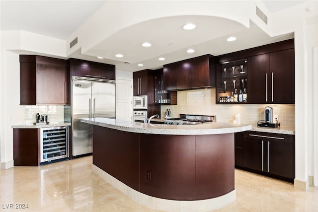 kitchen with an island with sink, custom exhaust hood, built in appliances, wine cooler, and decorative backsplash