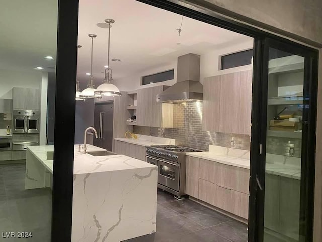 kitchen with light stone counters, stainless steel appliances, wall chimney range hood, sink, and an island with sink