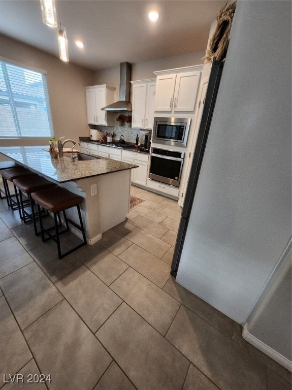 kitchen with wall chimney range hood, a center island with sink, a breakfast bar area, white cabinetry, and stainless steel appliances