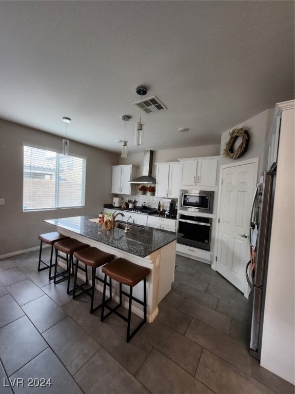 kitchen featuring a kitchen island with sink, wall chimney exhaust hood, stainless steel appliances, decorative light fixtures, and white cabinetry