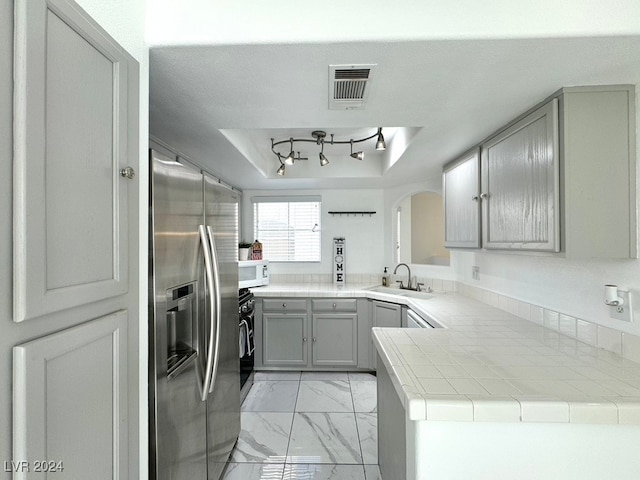 kitchen with tile counters, kitchen peninsula, gray cabinetry, stainless steel appliances, and sink