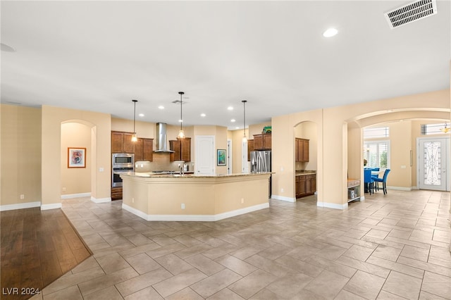 kitchen with a large island with sink, appliances with stainless steel finishes, tasteful backsplash, wall chimney exhaust hood, and hanging light fixtures