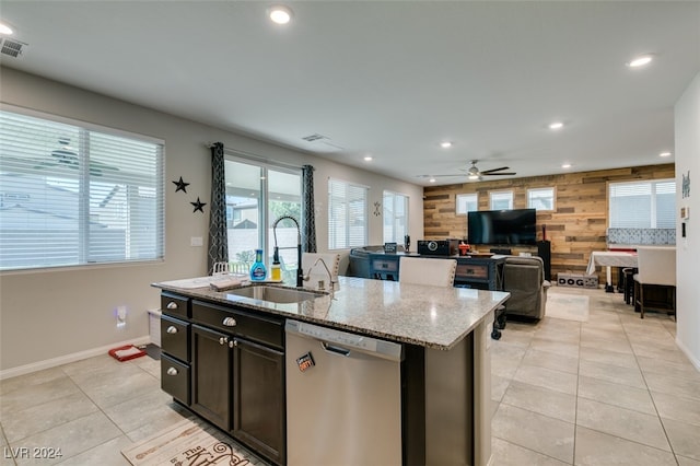 kitchen with ceiling fan, an island with sink, stainless steel dishwasher, sink, and wooden walls