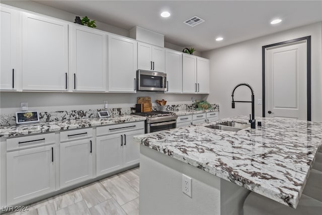 kitchen with sink, an island with sink, white cabinetry, stainless steel appliances, and light stone counters