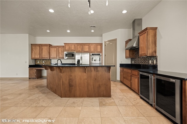 kitchen featuring wall chimney exhaust hood, stainless steel appliances, beverage cooler, and an island with sink