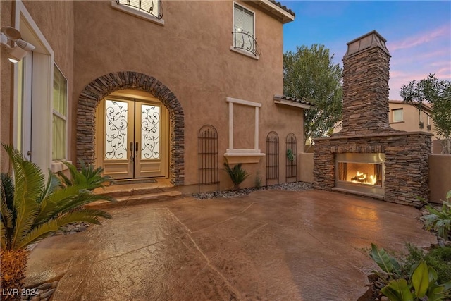 exterior entry at dusk with french doors, a patio, and an outdoor stone fireplace