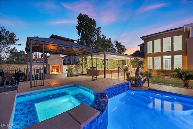 pool at dusk featuring an in ground hot tub, a gazebo, a patio, and exterior fireplace