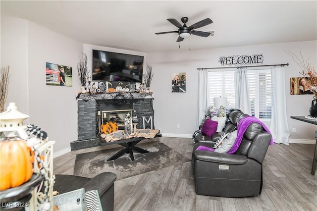 living area with a ceiling fan, baseboards, a stone fireplace, and wood finished floors