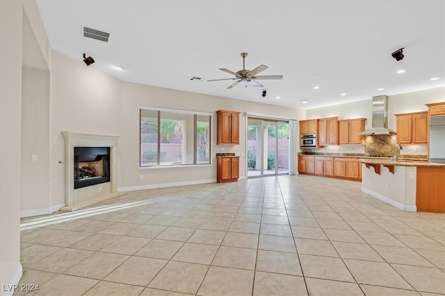 kitchen featuring wall chimney range hood, a breakfast bar area, ceiling fan, backsplash, and light tile patterned floors