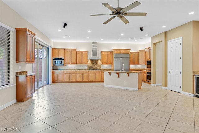 kitchen featuring ceiling fan, stainless steel appliances, wall chimney exhaust hood, a breakfast bar area, and a kitchen island with sink