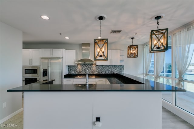 kitchen featuring wall chimney range hood, pendant lighting, white cabinets, built in appliances, and decorative backsplash