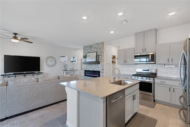 kitchen featuring an island with sink, sink, appliances with stainless steel finishes, light stone counters, and ceiling fan