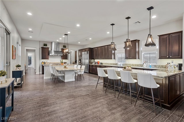 kitchen featuring wall chimney range hood, a spacious island, dark wood-type flooring, built in fridge, and decorative light fixtures