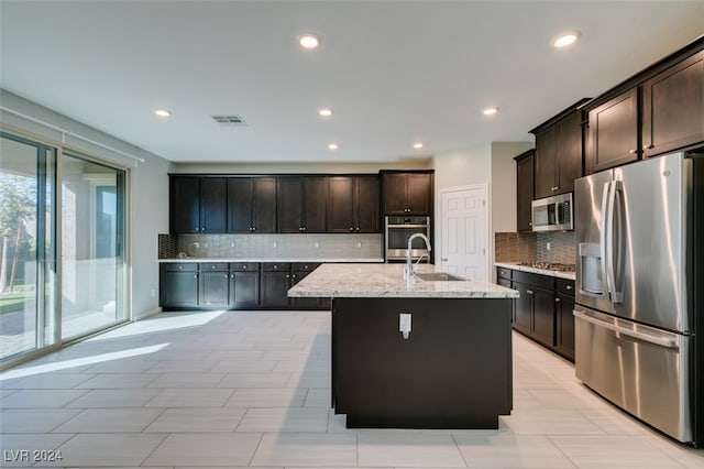 kitchen with sink, a kitchen island with sink, stainless steel appliances, dark brown cabinets, and light stone counters