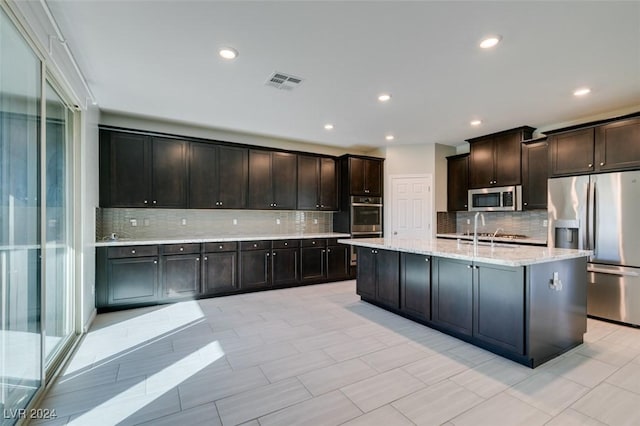 kitchen featuring a center island with sink, stainless steel appliances, decorative backsplash, dark brown cabinetry, and sink