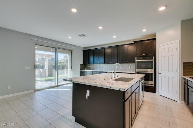 kitchen featuring a center island with sink, decorative backsplash, light stone countertops, double oven, and sink