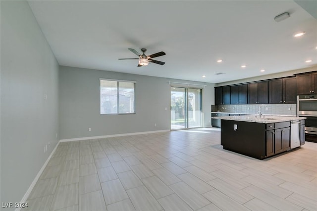 kitchen featuring stainless steel double oven, ceiling fan, decorative backsplash, a kitchen island with sink, and light stone counters