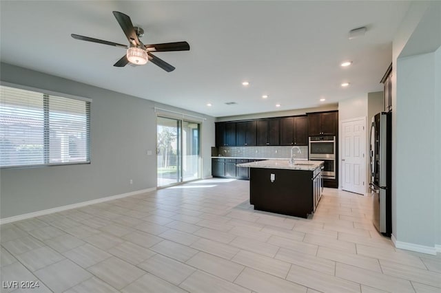 kitchen featuring tasteful backsplash, ceiling fan, a center island with sink, appliances with stainless steel finishes, and light stone counters