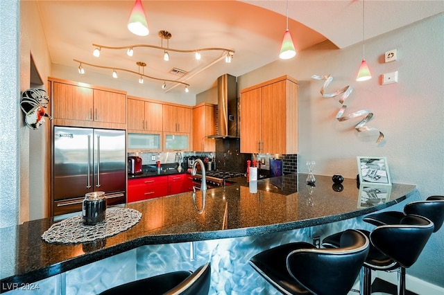 kitchen with wall chimney range hood, a kitchen bar, kitchen peninsula, stainless steel built in refrigerator, and pendant lighting