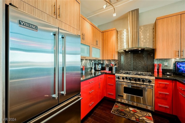 kitchen with premium appliances, wall chimney range hood, dark wood-type flooring, dark stone countertops, and decorative backsplash