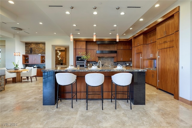 kitchen with a large island with sink, decorative backsplash, dark stone counters, a kitchen bar, and decorative light fixtures