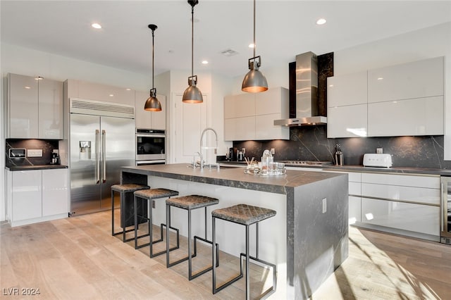 kitchen featuring wall chimney range hood, appliances with stainless steel finishes, light wood-type flooring, pendant lighting, and a center island with sink