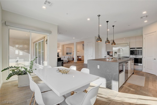 kitchen with sink, an island with sink, hanging light fixtures, light hardwood / wood-style floors, and stainless steel appliances