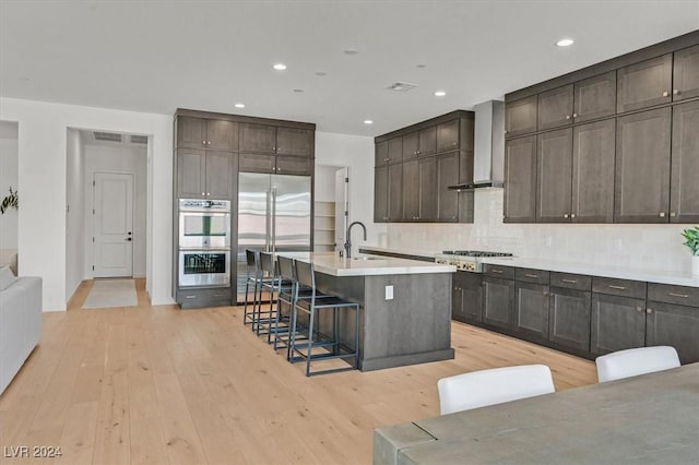 kitchen featuring wall chimney exhaust hood, a kitchen breakfast bar, a kitchen island with sink, appliances with stainless steel finishes, and light wood-type flooring