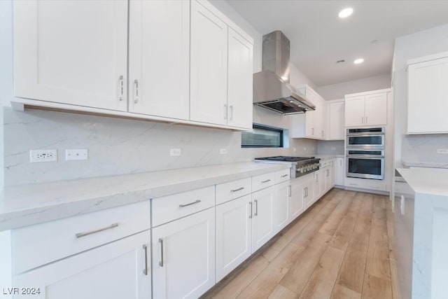 kitchen with light stone counters, white cabinetry, light hardwood / wood-style floors, wall chimney exhaust hood, and stainless steel appliances