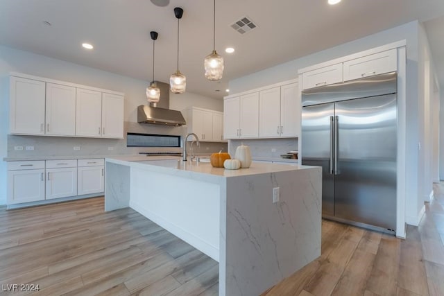 kitchen with stainless steel built in refrigerator, wall chimney range hood, decorative light fixtures, white cabinets, and light hardwood / wood-style floors