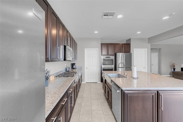 kitchen featuring light stone countertops, a kitchen island with sink, dark brown cabinets, light tile patterned floors, and appliances with stainless steel finishes