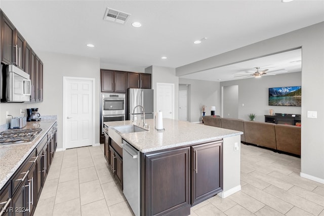 kitchen featuring a kitchen island with sink, ceiling fan, light stone countertops, dark brown cabinets, and stainless steel appliances