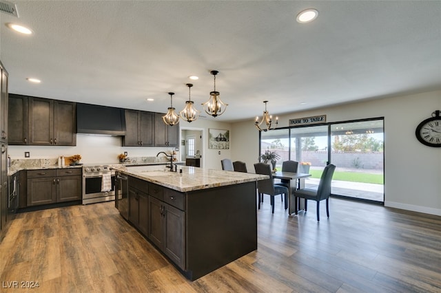 kitchen featuring wall chimney range hood, a kitchen island with sink, dark hardwood / wood-style floors, stainless steel appliances, and decorative light fixtures