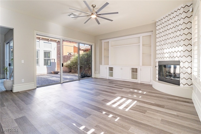 unfurnished living room with a large fireplace, built in features, light wood-type flooring, and ceiling fan