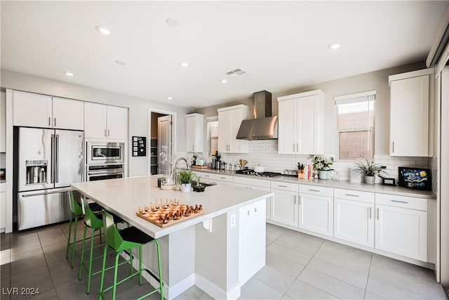 kitchen with wall chimney range hood, white cabinetry, a kitchen island with sink, and appliances with stainless steel finishes