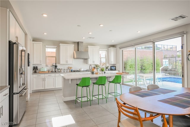 kitchen featuring a wealth of natural light, stainless steel fridge with ice dispenser, white cabinetry, and wall chimney exhaust hood