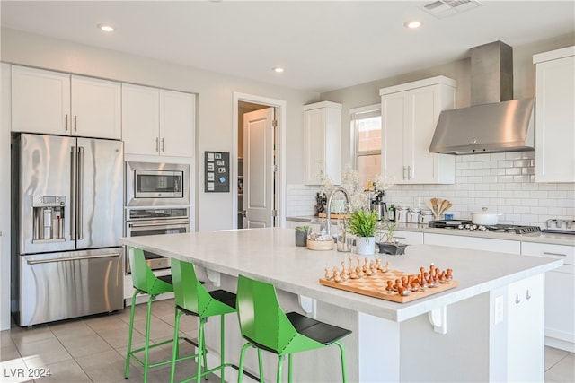 kitchen with a center island with sink, appliances with stainless steel finishes, a kitchen breakfast bar, white cabinets, and wall chimney range hood