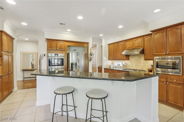 kitchen with stainless steel appliances, dark stone countertops, a center island with sink, ornamental molding, and light tile patterned floors