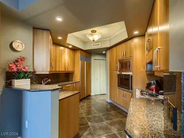 kitchen with built in appliances, backsplash, a tray ceiling, stone counters, and dark tile patterned flooring