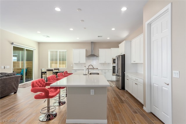 kitchen featuring hardwood / wood-style floors, a kitchen island with sink, wall chimney range hood, stainless steel refrigerator, and white cabinetry