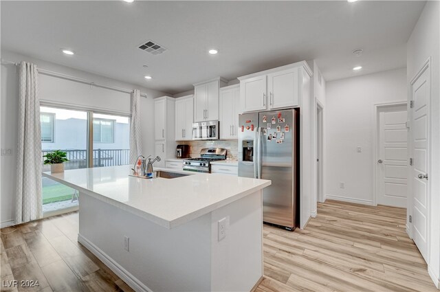 kitchen featuring light wood-type flooring, appliances with stainless steel finishes, decorative backsplash, an island with sink, and white cabinets