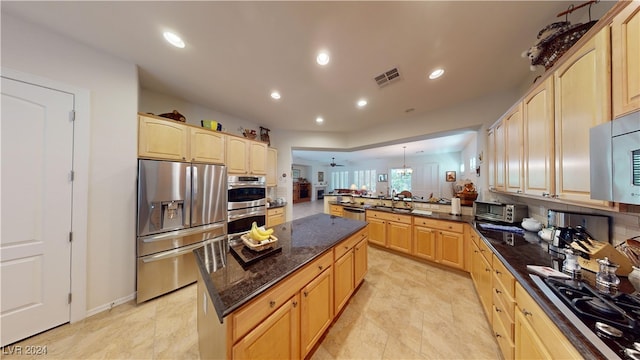 kitchen with kitchen peninsula, light brown cabinets, pendant lighting, appliances with stainless steel finishes, and dark stone countertops