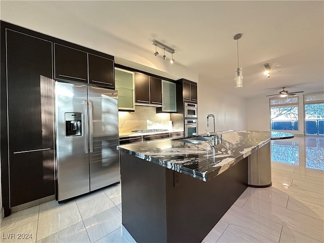 kitchen featuring a center island with sink, decorative backsplash, appliances with stainless steel finishes, decorative light fixtures, and dark stone countertops