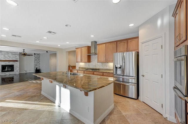 kitchen with wall chimney range hood, sink, stainless steel appliances, a fireplace, and dark stone counters