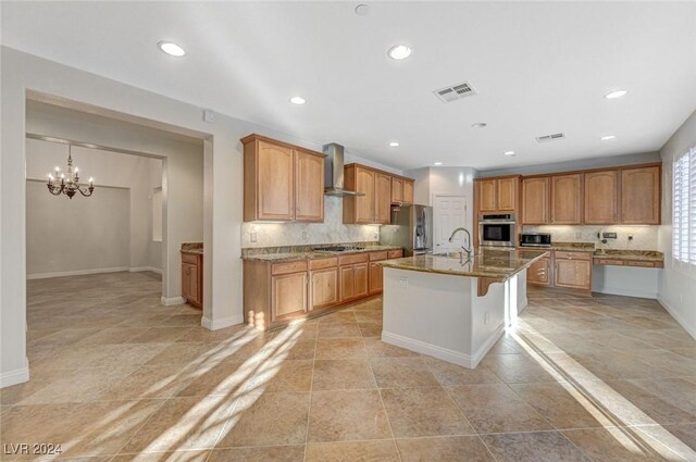 kitchen with stainless steel appliances, decorative backsplash, a chandelier, a kitchen island with sink, and wall chimney range hood