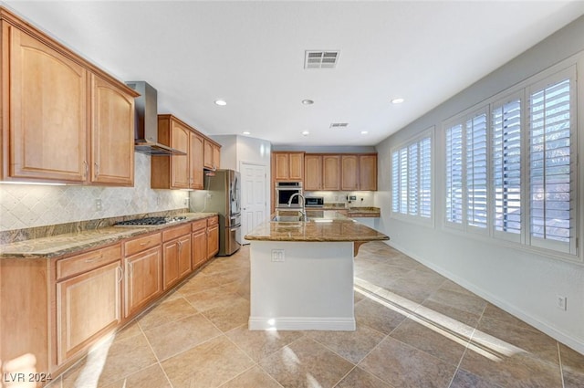 kitchen featuring wall chimney exhaust hood, an island with sink, stone counters, stainless steel appliances, and decorative backsplash